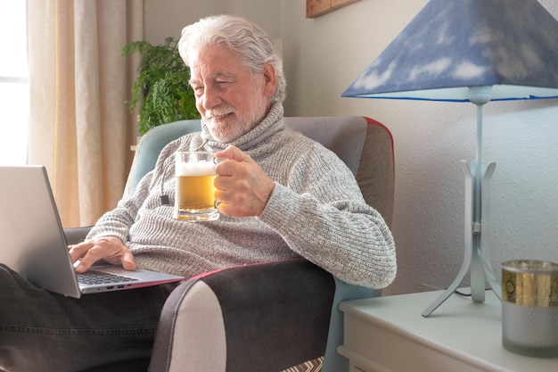 Starszy kaukaski mężczyzna relaksuje się w domu na fotelu przeglądając laptopa, trzymając piwo w kolorze blond