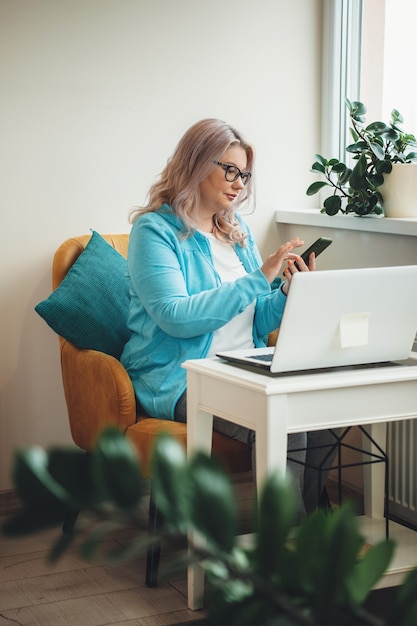 Starszy kobieta o blond włosach i okularach pracuje na telefonie i laptopie w domu podczas kwarantanny