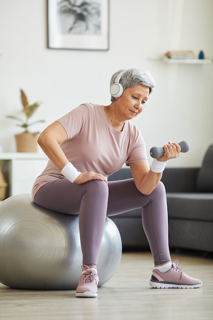 Starszy kobieta w słuchawkach słuchanie muzyki i ćwiczenia z hantlami na piłce fitness w salonie w domu