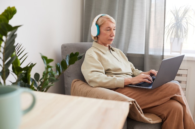 Starszy kobieta w słuchawkach za pomocą laptopa, odpoczywając na fotelu w domu