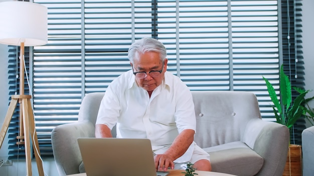 Starszy Mężczyzna Czytający Wiadomości Z Tabletem Na Kanapie W Domu. Siedzący W Salonie Starszy Azjata Szuka Informacji W Internecie
