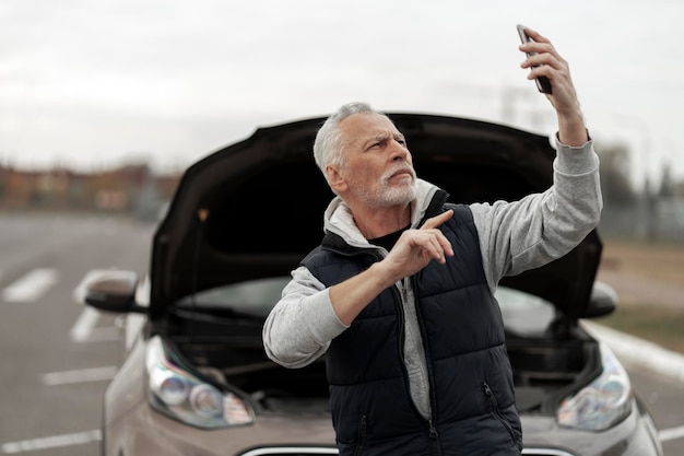 Starszy mężczyzna kierowca rozmawia na smartfonie w pobliżu zepsutego samochodu z otwartą maską Koncepcja ubezpieczenia samochodu