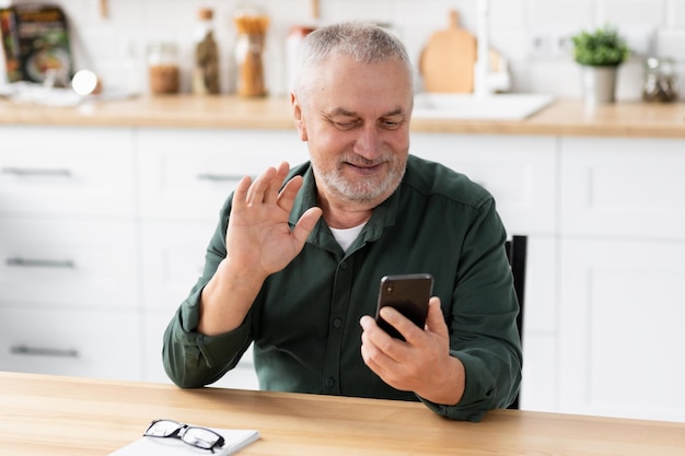 Starszy mężczyzna komunikuje się online za pomocą telefonu komórkowego, siedząc w domu w kuchni. Starszy mężczyzna trzymający smartfon w rękach rozmawiający podczas rozmowy wideo