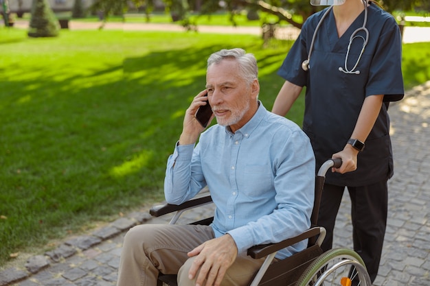 Starszy mężczyzna odzyskujący pacjenta na wózku inwalidzkim rozmawiający przez telefon podczas spaceru z pielęgniarką w