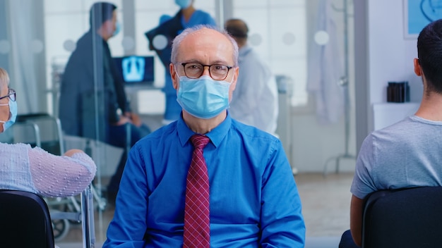 Starszy mężczyzna patrząc na kamerę nosząc maskę przeciwko koronawirusowi w poczekalni szpitala. Niepełnosprawna stara kobieta z balkonikiem w klinice do badania