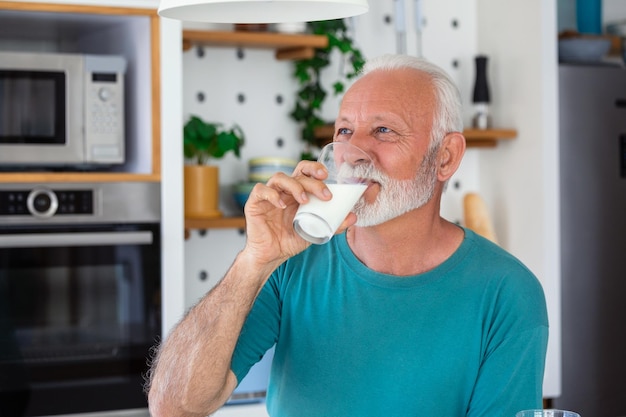 Starszy mężczyzna pijący szklankę mleka z szczęśliwą twarzą stojący i uśmiechnięty Przystojny starszy mężczyzna pijący szklankę świeżego mleka w kuchni