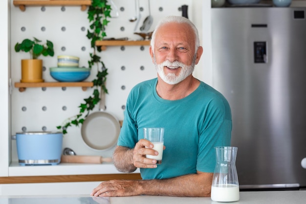 Starszy mężczyzna pijący szklankę mleka z szczęśliwą twarzą stojący i uśmiechnięty Przystojny starszy mężczyzna pijący szklankę świeżego mleka w kuchni