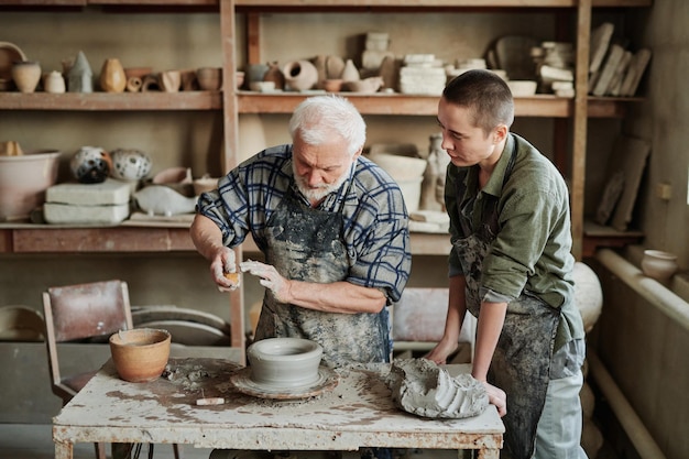 Starszy mężczyzna pokazujący swojemu asystentowi w warsztacie garncarskim proces tworzenia ceramicznych waz