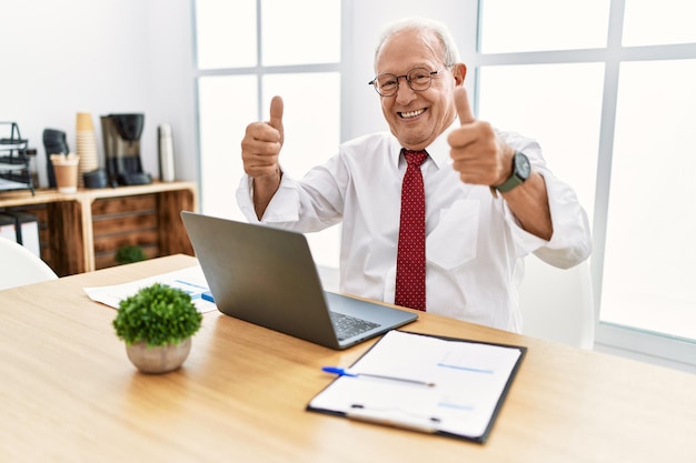 Starszy mężczyzna pracujący w biurze za pomocą laptopa komputerowego zatwierdzający pozytywny gest ręką, uśmiechnięty i szczęśliwy z powodu sukcesu. gest zwycięzcy.