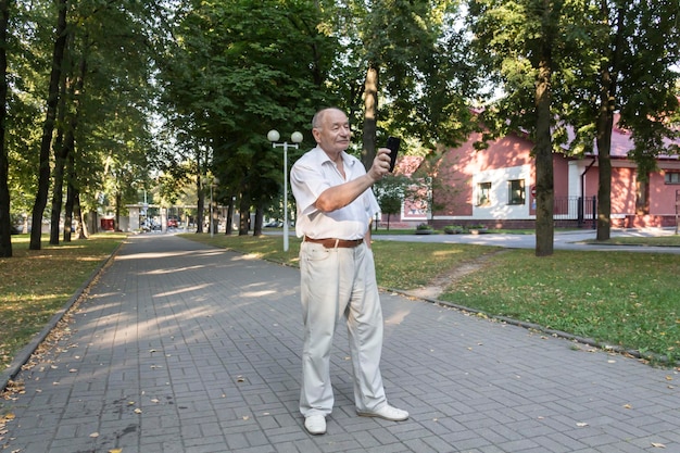 Starszy mężczyzna spaceruje samotnie po parku latem Nowoczesny emeryt, biznesmen w białej koszuli i spodniach robi zdjęcia aparatem w telefonie komórkowym