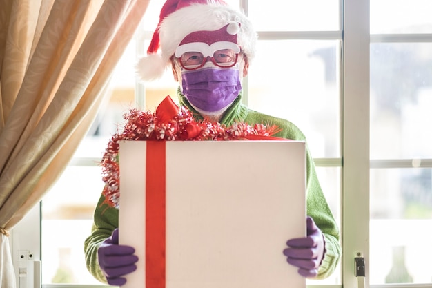 Zdjęcie starszy mężczyzna w czapce mikołaja w masce chirurgicznej, trzymający świąteczne pudełko z prezentem do dostawy do domu