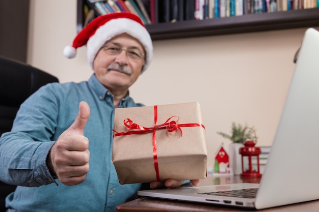 Starszy mężczyzna w czapce Świętego Mikołaja Wręcza prezent i rozmawia z przyjaciółmi i dziećmi za pomocą laptopa. Boże Narodzenie w okresie koronawirusa.