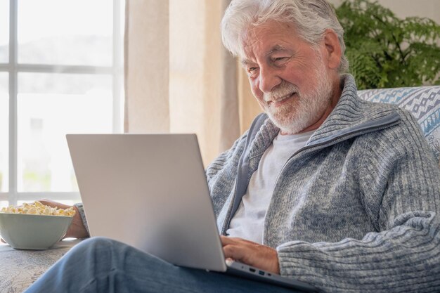 Starszy mężczyzna z lat 70. siedzący na kanapie przeglądający laptopa starszy patrzący na ekran laptopa surfujący po sieci starsza generacja i nowoczesna aplikacja technologiczna koncepcja łatwego użytkowania