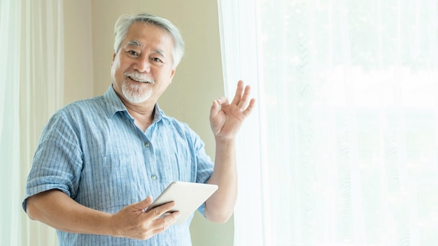 Starszy mężczyzna za pomocą smartfona i pokazuje znak ok z rękami tablet komputer uśmiechnięty czuje się szczęśliwy w sypialni w domu styl życia koncepcja seniora