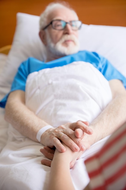 Starszy pacjent w łóżku szpitalnym trzymający się za ręce z gościem