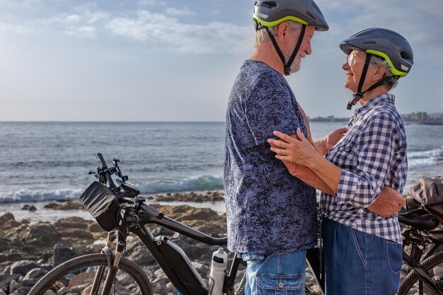 Starszy para kaukaski obejmując się przed morzem podczas jazdy poza drogą na kamienistej plaży z rowerami o zachodzie słońca Autentyczna koncepcja życia starszych emerytów