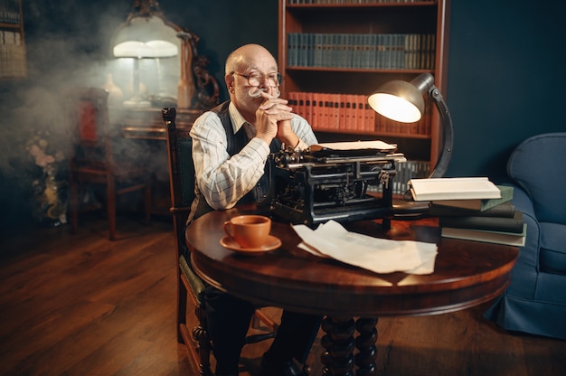 Zdjęcie starszy pisarz myśli o zabytkowej maszynie do pisania w domowym biurze. stary człowiek w okularach pisze powieść literacką w pokoju z dymem, inspiracją