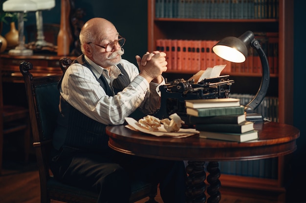 Starszy Pisarz Myśli O Zabytkowej Maszynie Do Pisania W Domowym Biurze. Stary Człowiek W Okularach Pisze Powieść Literacką W Pokoju Z Dymem