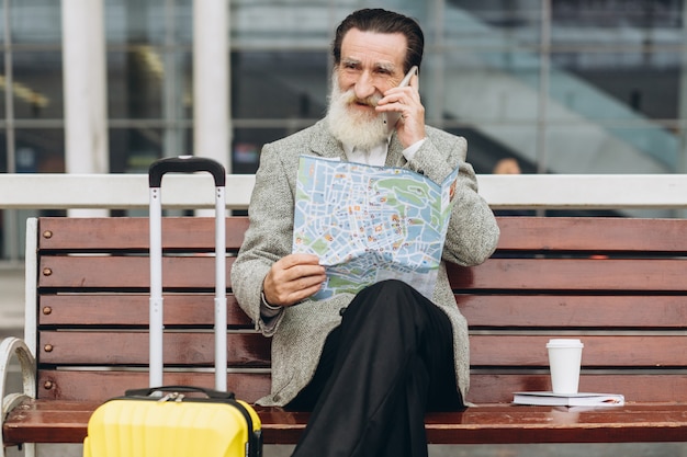 Starszy Siwobrody Mężczyzna Siedzi Na ławce Z Walizką, Patrzy Na Mapę Miasta I Telefon W Budynku Lotniska