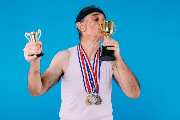 Zdjęcie starszy sportowiec ze śladami słońca na rękach, z trzema medalami na szyi całuje trofeum, na niebieskim tle