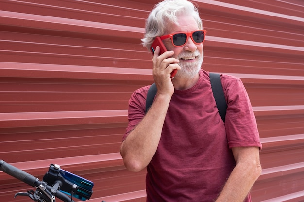 Starszy uśmiechnięty rowerzysta w wycieczce plenerowej odpoczywający na czerwonym metalowym panelu za pomocą telefonu komórkowego
