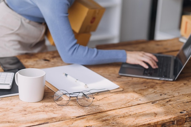 Startup MŚP mały przedsiębiorca niezależnej azjatyckiej kobiety noszącej fartuch za pomocą laptopa i pudełka do otrzymywania i przeglądania zamówień online w celu przygotowania się do pakowania sprzedaży klientom pomysłów biznesowych MŚP online