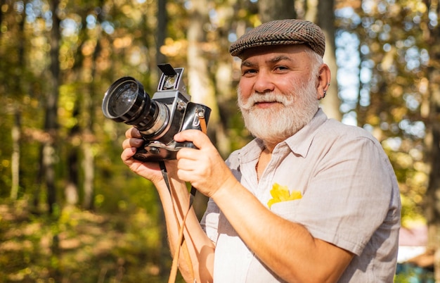 Stary fotograf używa aparatu retro Starszy mężczyzna relaksuje się w letniej przyrodzie Brodaty mężczyzna trzyma aparat retro i robi zdjęcie światła słonecznego w okresie letnim fotograf fotograf trzyma aparat w przyrodzie