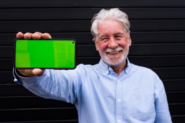 Stary i dojrzały mężczyzna trzymający smartfon pokazujący go przed kamerą z zielonym ekranem uśmiechający się szczęśliwy portret dorosłego za pomocą technologii