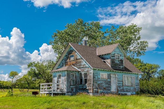 Stary, opuszczony dom wiejski z drzewami, trawą i błękitnym niebem w Kayville, Saskatchewan, Kanada