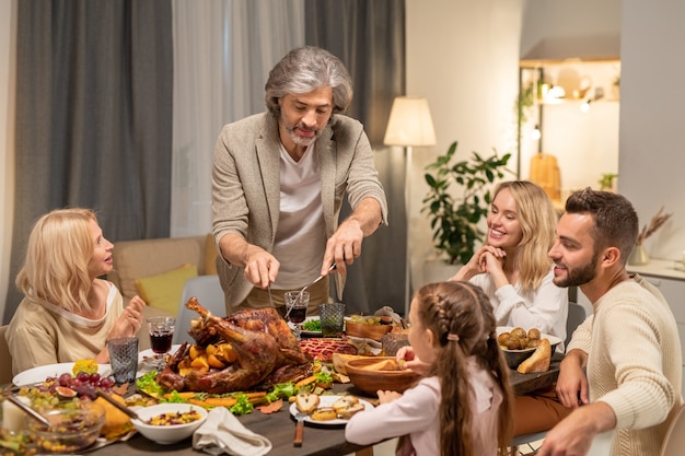 Starzejący się mężczyzna z nożem i widelcem kroi domowej roboty pieczonego indyka na środku świątecznego stołu, podczas gdy jego rodzina patrzy na niego przy kolacji