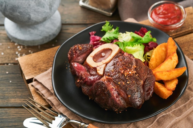 Steki Osso Buco Plasterki wołowiny z grilla Osso Buco lub porterhouse Stek mięsny z przyozdobionym sałatką i frytkami na czarnej marmurowej desce na starym drewnianym tle