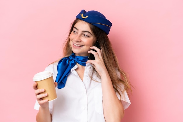 Stewardesa samolotu kaukaska kobieta na białym tle na różowym tle trzymająca kawę na wynos i telefon komórkowy