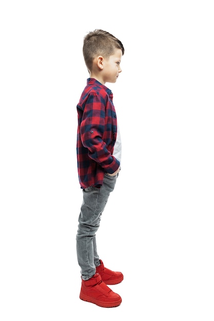 Stoi chłopiec w wieku 7 lat w dżinsach i czerwonej koszuli w kratę. Pełna wysokość. Widok profilu. Na białym tle na białej ścianie. Pionowy.
