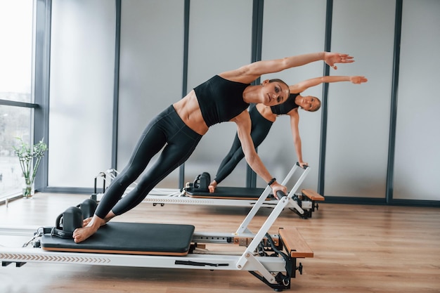Stojąc na sprzęcie gimnastycznym i rozciągając się Dwie kobiety w sportowym stroju i szczupłych ciałach spędzają razem dzień jogi fitness w pomieszczeniu