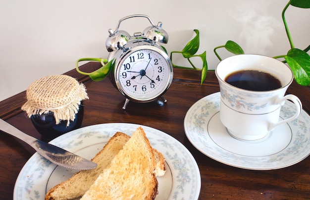 Zdjęcie stół śniadaniowy z herbatą lub kawą w delikatnej filiżance i spodeczku z kromkami tostów i garnkiem dżemu oraz retro budzikiem