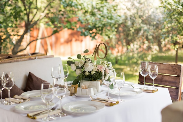 Stół weselny ozdobiony świeżymi kwiatami w mosiężnym wazonie. Florystyka ślubna. Stół bankietowy dla gości na zewnątrz z widokiem na zieloną przyrodę. Bukiet z różami, eustomą i liśćmi eukaliptusa