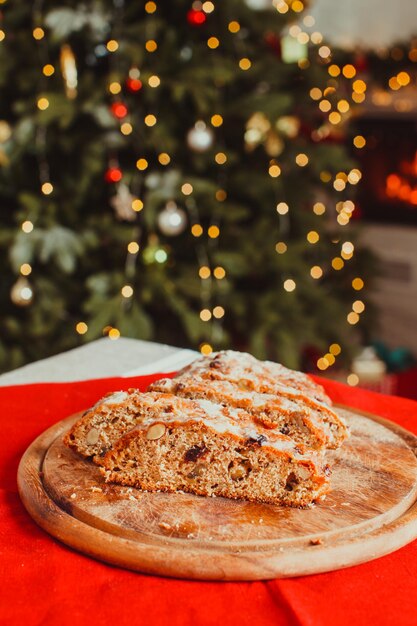 Stollen Boże Narodzenie. Tradycyjny bochenek ze słodkich owoców z cukrem pudrem. Świąteczny stół świąteczny ze stollen. Choinka i płonący kominek w pokoju w tle