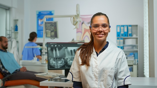 Stomatolog lekarz uśmiecha się do kamery w gabinecie stomatologicznym, podczas gdy asystent przygotowuje człowieka do chirurgii stomatologicznej w tle. Dentysta kobieta patrząc na kamerę internetową siedzącą na krześle w pokoju stomatologicznym