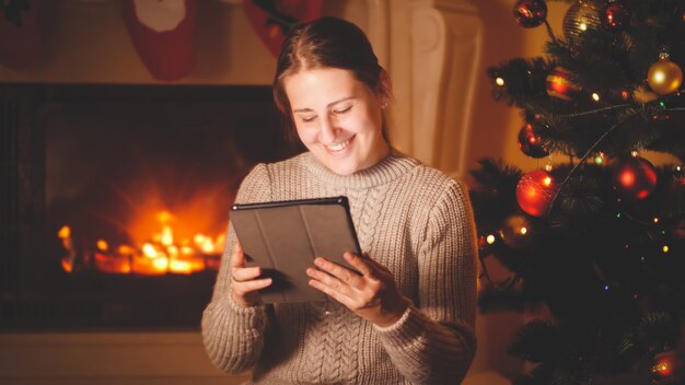 Stonowany portret szczęśliwej uśmiechniętej kobiety trzymającej i używającej cyfrowego tabletu w Wigilię Bożego Narodzenia przy kominku