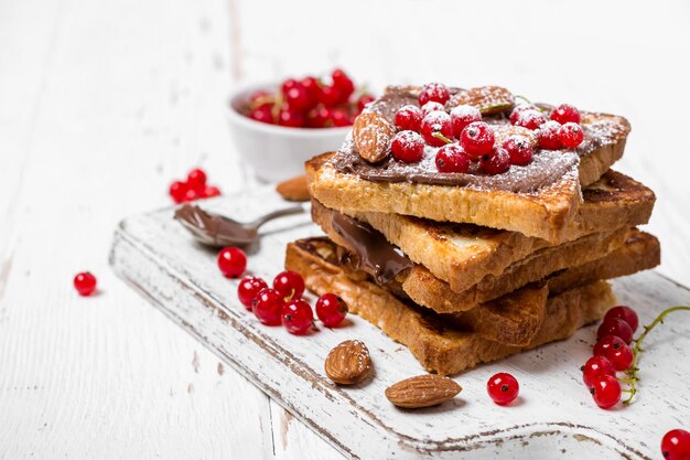 Stos smażonych tostów z kremem czekoladowym, czerwoną porzeczką i migdałami na białym drewnianym tle