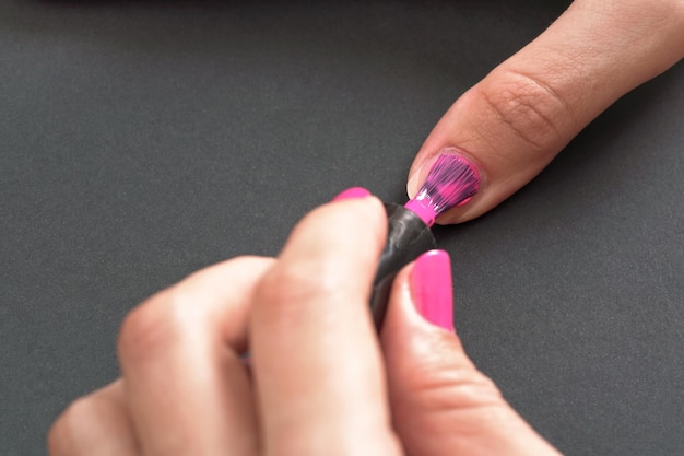 Stosując różowy lakier do paznokci - zbliżenie zdjęcie rąk kobiety, mały pędzel i paznokcie na szarym czarnym tle.