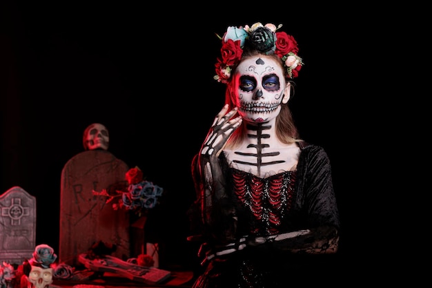 Straszna bogini zmarłych w czarnym kostiumie i tatuażu na czaszce Catriny, nosząca kwiatową opaskę z różami. Świętujemy meksykańską tradycję dios de los muertos na halloween w studio.