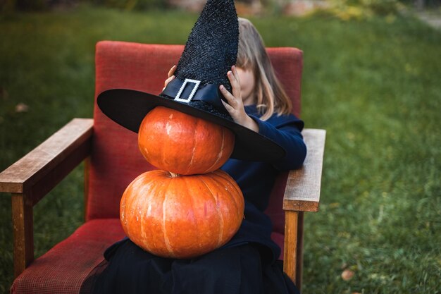 Straszna mała dziewczynka w kapeluszu czarownicy z wieloma dyniami świętuje halloweenowe wakacje Siedząc na fotelu w płaszczu z dynią Stylowy obraz Horror zabawa na imprezie dla dzieci w stodole na ulicy