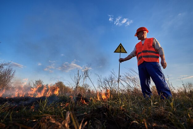 Strażak-ekolog gaszący ogień na polu wieczorem Mężczyzna w pomarańczowej kamizelce i hełmie w pobliżu płonącej trawy z dymem trzymający znak ostrzegawczy z znakiem wykrzyku Koncepcja katastrofy naturalnej