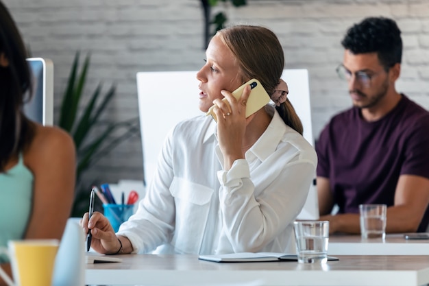 Strzał inteligentnej bizneswoman rozmawia przez telefon komórkowy podczas pracy z komputerem w biurze.