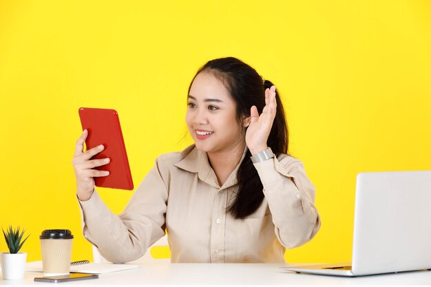 Strzał studio Azjatycka pulchna pulchna sekretarka pracownik pracownika siedzącego trzymając tablet z ekranem dotykowym pozdrowienie macha ręką powiedzieć cześć cześć do widzenia z klientem klienta na rozmowę wideo na żółtym tle.