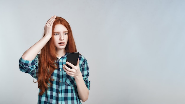 Zdjęcie strzał studio zszokowanej rudej kobiety w casual, trzymając rękę na głowie i patrząc na telefon komórkowy, stojąc na szarym tle. negatywne emocje. cyfrowy