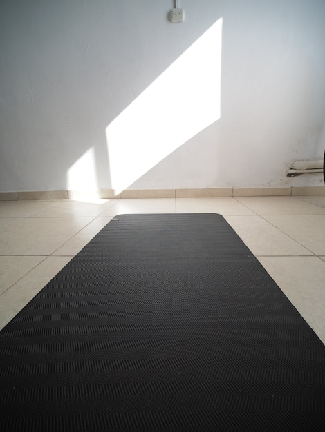Zdjęcie strzał w pov. rozwinięta czarna mata do jogi na podłodze w pokoju, światło słoneczne na białej ścianie.