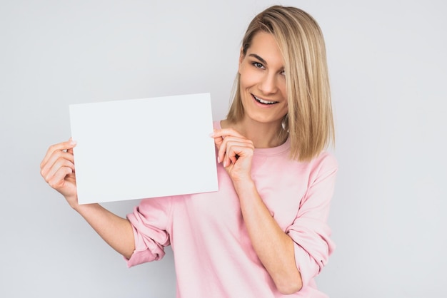 Zdjęcie strzał zbliżenie piękny szczęśliwy uśmiechający się blond kobieta sobie różowy sweter pozowanie z białego pustego papieru z miejsca na kopię informacji o reklamie i patrząc na kamery na białej ścianie