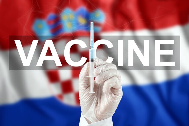 Strzykawka ze szczepionką w dłoni lekarza na tle chorwackiej flagi. Koronawirus COVID-19, ochrona immunologiczna, leczenie przeciwko zakażeniu nCoV 2019, szczepienia.